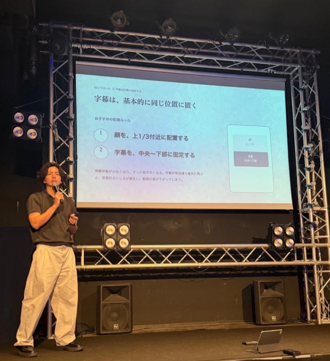 ステージ上で講演する代表 新屋敷。スクリーンには動画の字幕配置ルールを解説するスライド